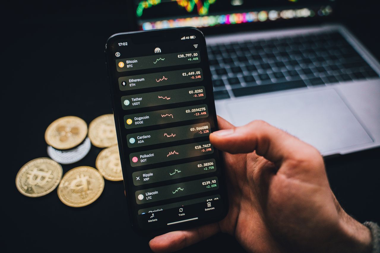 our-experience A person checks cryptocurrency values on a smartphone with a laptop and Bitcoins nearby.